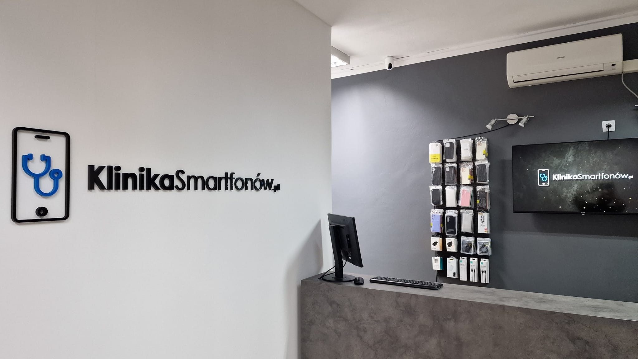 Recepcja serwisu telefonów z logo firmy 'KlinikaSmartfonow.pl' w formie telefonu ze stetoskopem, ekspozycją etui na telefony na ścianie i monitorem prezentującym logo firmy.