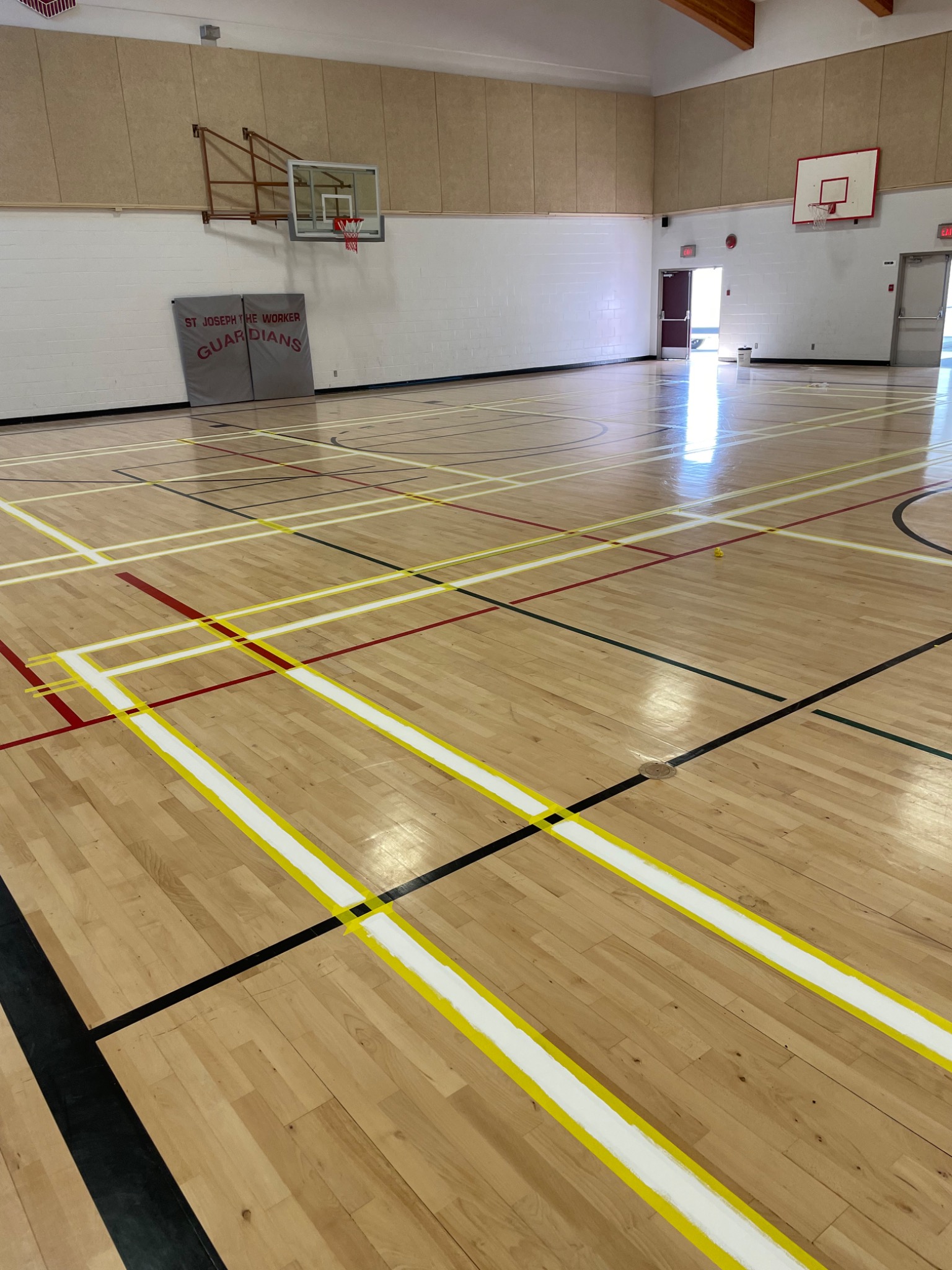 Sala gimnastyczna z drewnianą podłogą w trakcie malowania linii boiska, widoczne taśmy malarskie i świeżo nałożona biała farba.