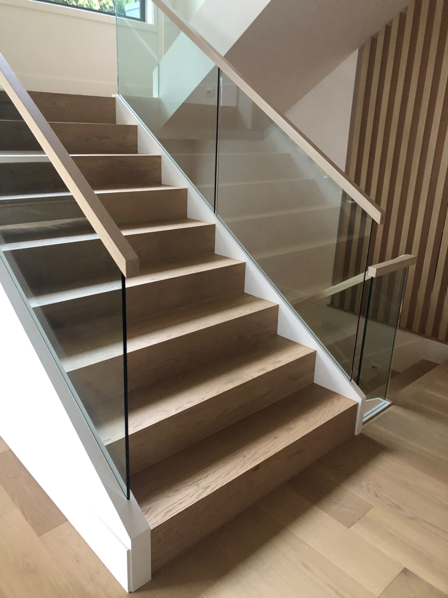 Nowoczesne, jasne schody z drewnianymi stopniami i szklaną balustradą, widoczne pionowe panele dekoracyjne na ścianie w tle.