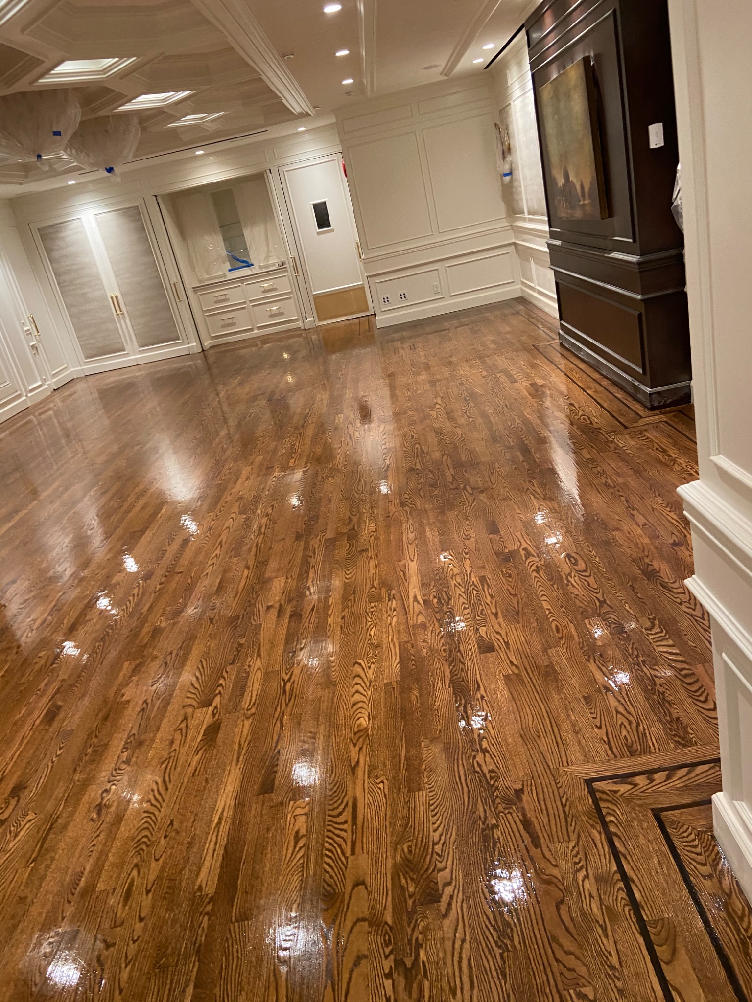 Świeżo polakierowany, błyszczący parkiet z wyraźnym usłojeniem, w przestronnym wnętrzu z białymi ścianami i wbudowanymi szafami, widoczne odbicia światła na powierzchni podłogi.