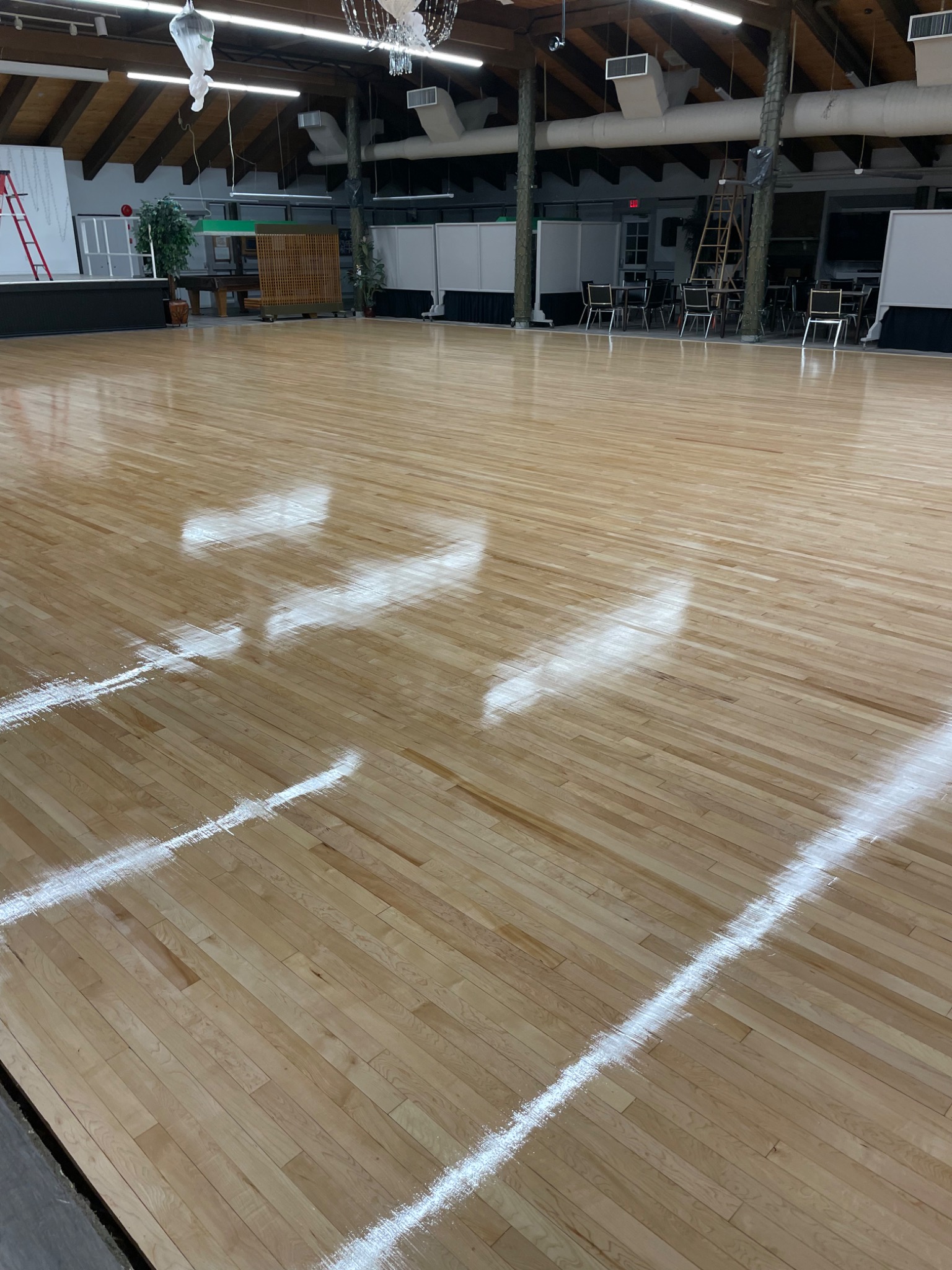 Błyszcząca, jasna podłoga parkietowa z widocznymi liniami wyznaczającymi obszar, prawdopodobnie świeżo polakierowana, w tle widoczne krzesła i drabina.