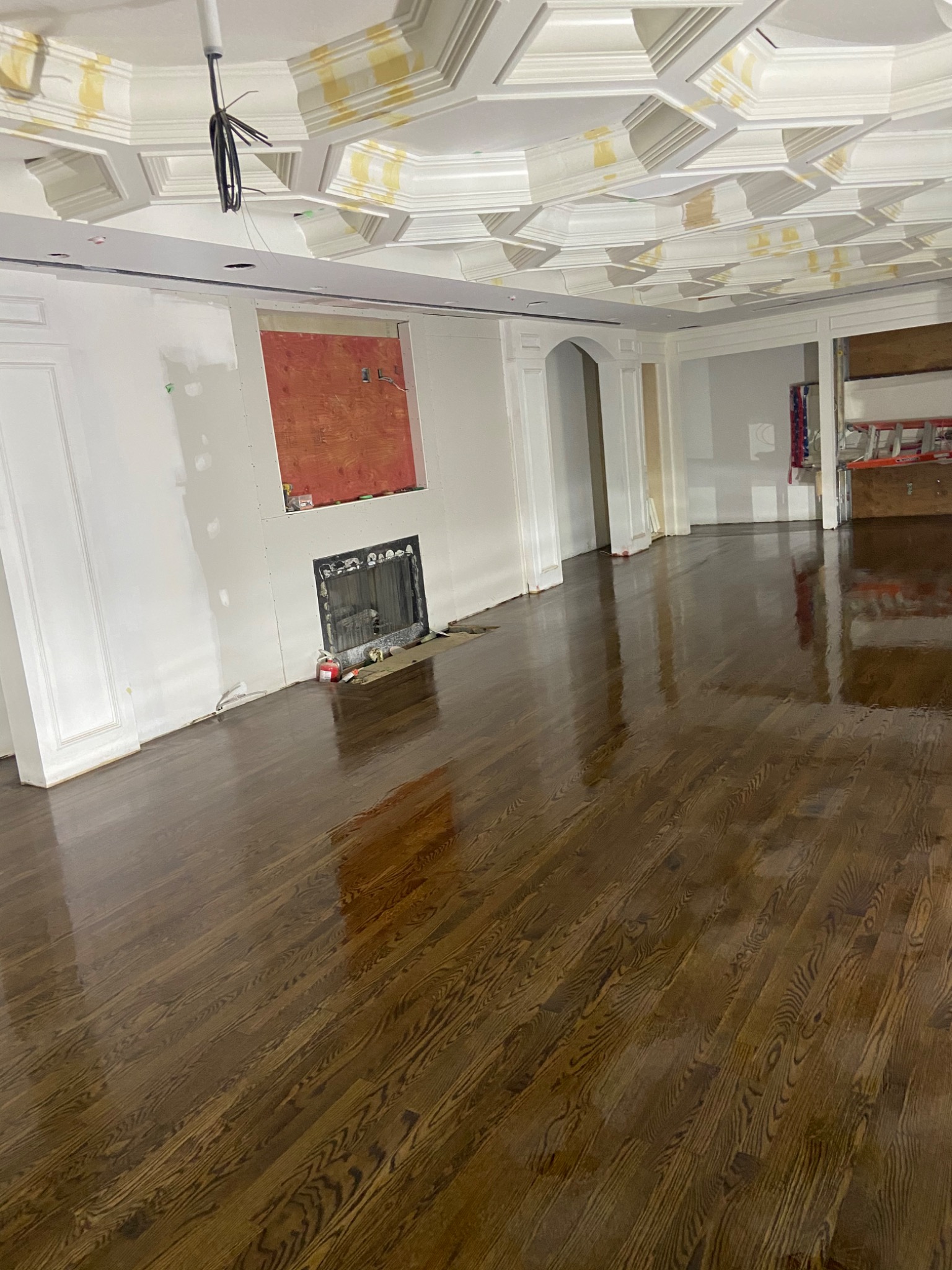 Błyszcząca, świeżo polakierowana podłoga z ciemnego drewna w przestronnym pokoju podczas remontu, widoczne zabezpieczenia na suficie, otwór na kominek i przygotowane ściany.