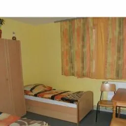 Pokój z pojedynczym łóżkiem, biurkiem, krzesłem i szafą w jasnych kolorach, z globusem na biurku i zasłonami w oknie. Doniczka z kwiatem stoi na szafie.