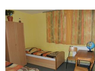 Pokój z pojedynczym łóżkiem, biurkiem, krzesłem i szafą w jasnych kolorach, z globusem na biurku i zasłonami w oknie. Doniczka z kwiatem stoi na szafie.