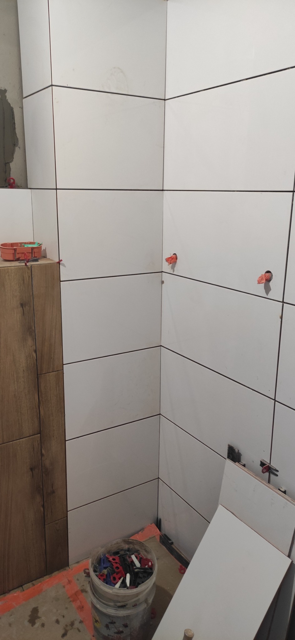 Białe, prostokątne płytki ceramiczne układane na ścianie w łazience, z widocznymi systemami poziomowania płytek, fragment ściany wyłożony płytkami imitującymi drewno, wiadro z klipsami...