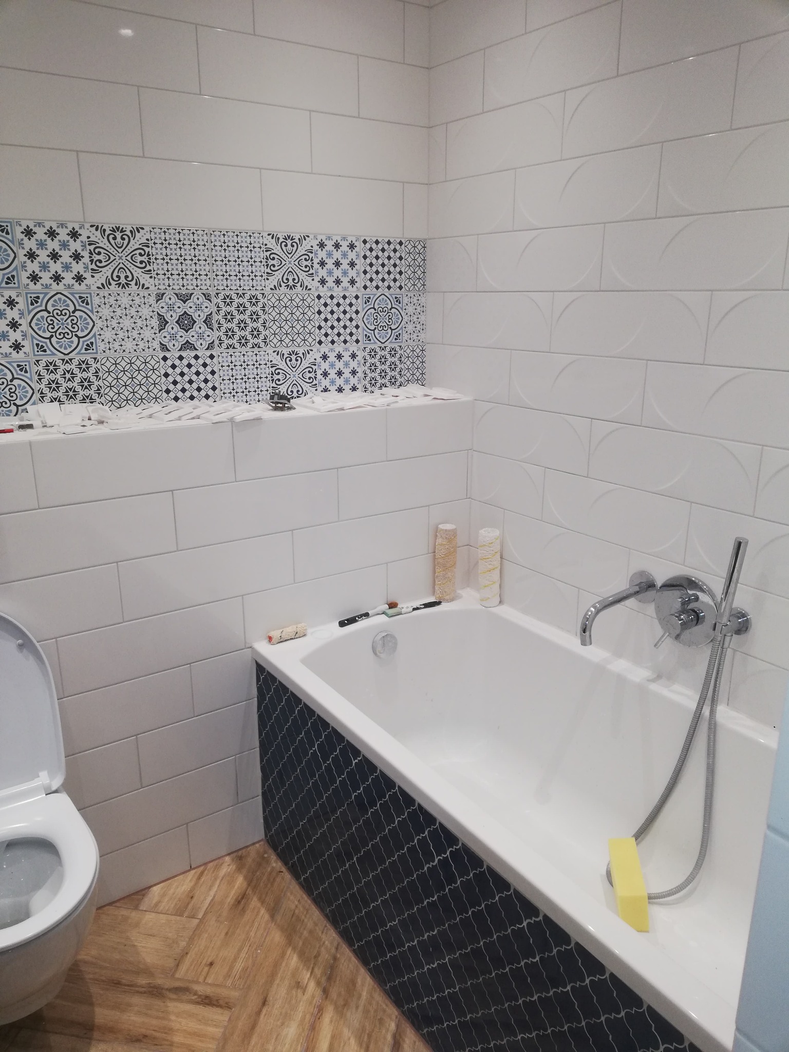 Niedokończona łazienka z białą wanną obudowaną mozaiką, białe płytki ścienne z dekoracyjnym pasem płytek w stylu marokańskim, drewnopodobna podłoga, widoczne narzędzia glazurnicze.