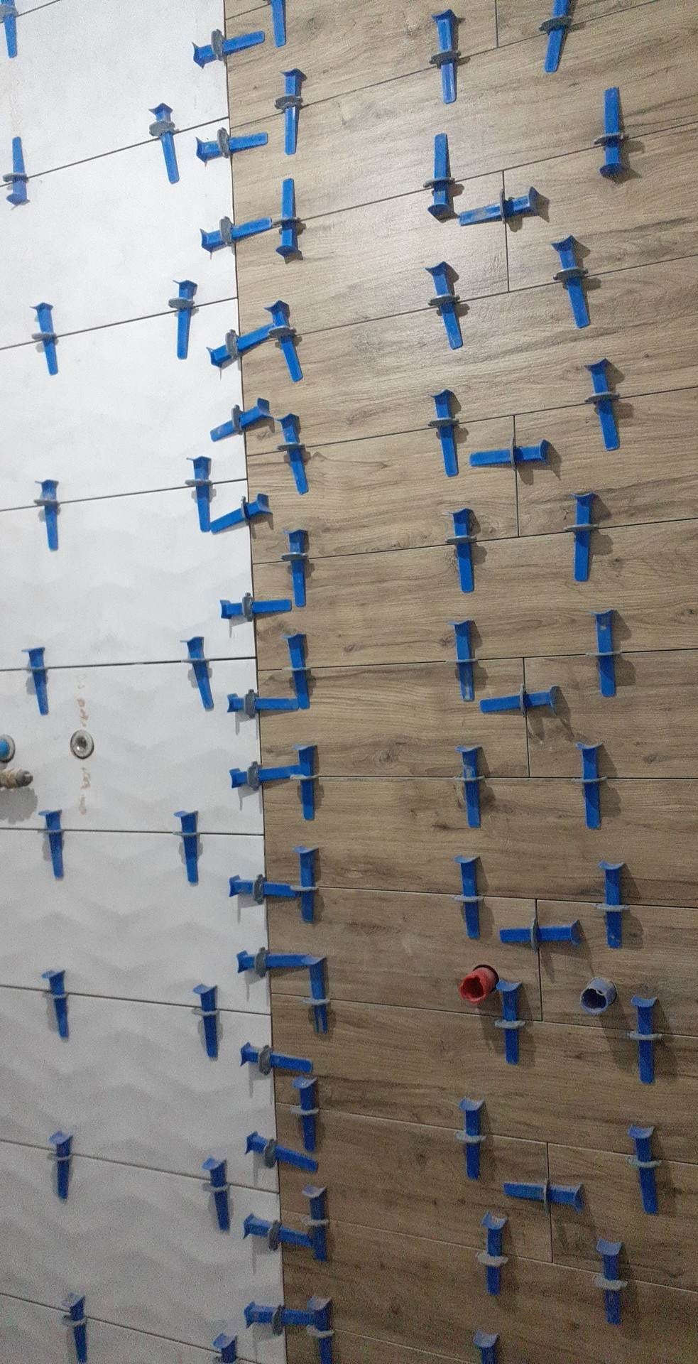 Układanie płytek imitujących drewno i białych płytek z użyciem niebieskich klipsów poziomujących, widoczne przygotowane otwory na instalacje.