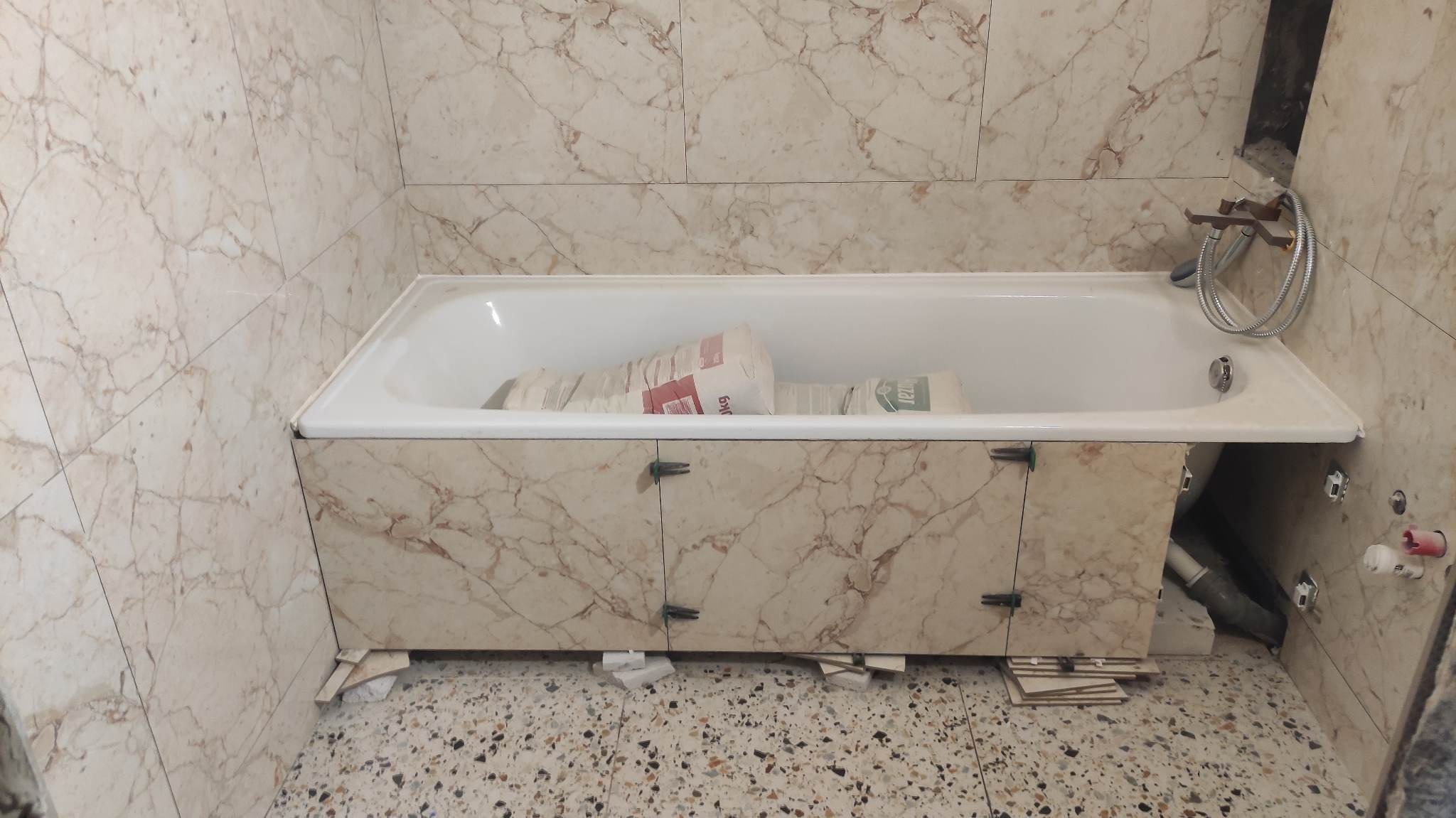 Wanna obudowana płytkami imitującymi marmur, widoczne worki z zaprawą wewnątrz wanny, płytki podłogowe w stylu lastryko i niedokończone prace wykończeniowe.