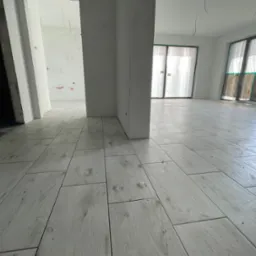 Układanie jasnych, podłużnych płytek podłogowych imitujących drewno w przestronnym, surowym wnętrzu. Widoczne niedociągnięcia i resztki po montażu.