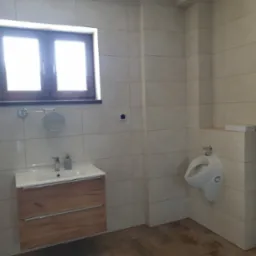 Wykończona łazienka z beżowymi płytkami, drewnianą szafką pod umywalką, białym pisuarze i oknem z widocznym światłem dziennym.