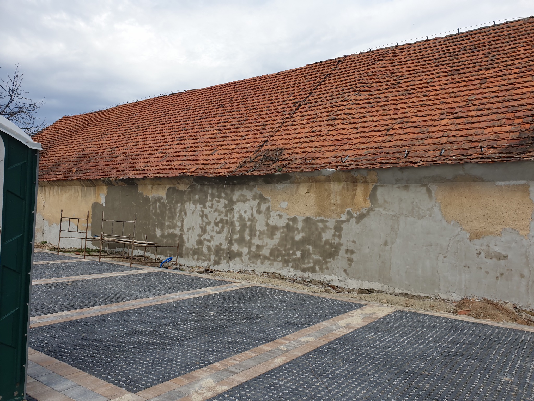 Ściana budynku z częściowo odnowionym tynkiem, widoczne rusztowanie i nowa nawierzchnia z kostki brukowej na podwórku, w tle czerwona dachówka.