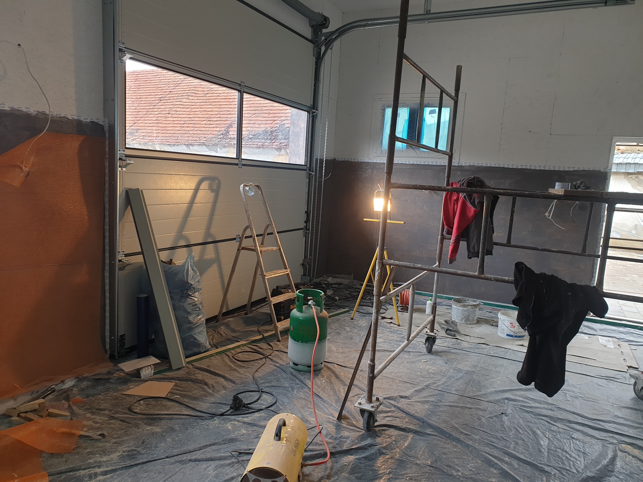 Wnętrze garażu w trakcie prac wykończeniowych: widoczne rusztowanie, butla gazowa, nagrzewnica, folia ochronna na podłodze, częściowo wykończone ściany z widoczną siatką i warstwą kleju.
