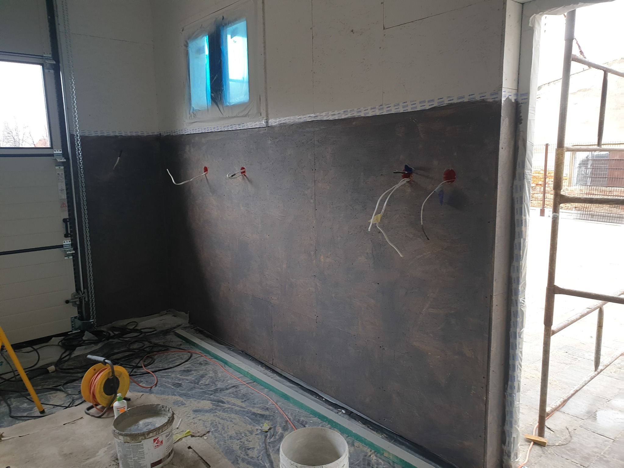 Wnętrze w trakcie remontu: częściowo otynkowana ściana z wystającymi przewodami elektrycznymi, okno zabezpieczone folią, widoczne narzędzia i wiadro z zaprawą.