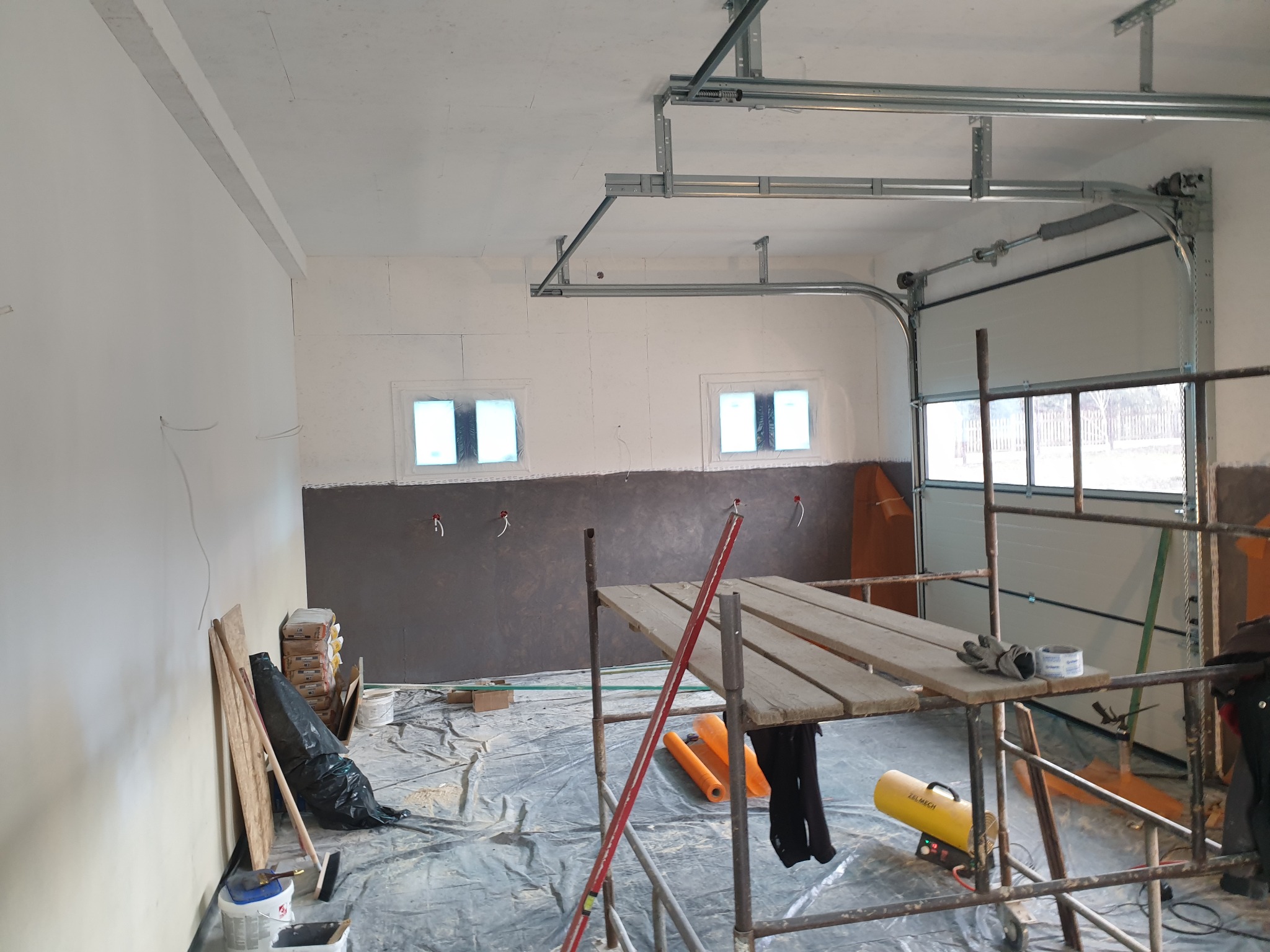 Wnętrze garażu w trakcie prac wykończeniowych: częściowo otynkowana ściana, rusztowanie z narzędziami, worki z zaprawą i folia ochronna na podłodze.