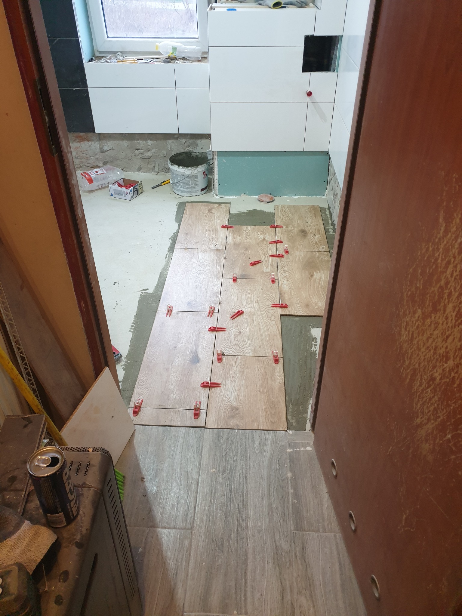 Układanie płytek podłogowych imitujących drewno w łazience, widoczne czerwone kliny poziomujące, wiadro z klejem i narzędzia w tle, drzwi po prawej stronie kadru.