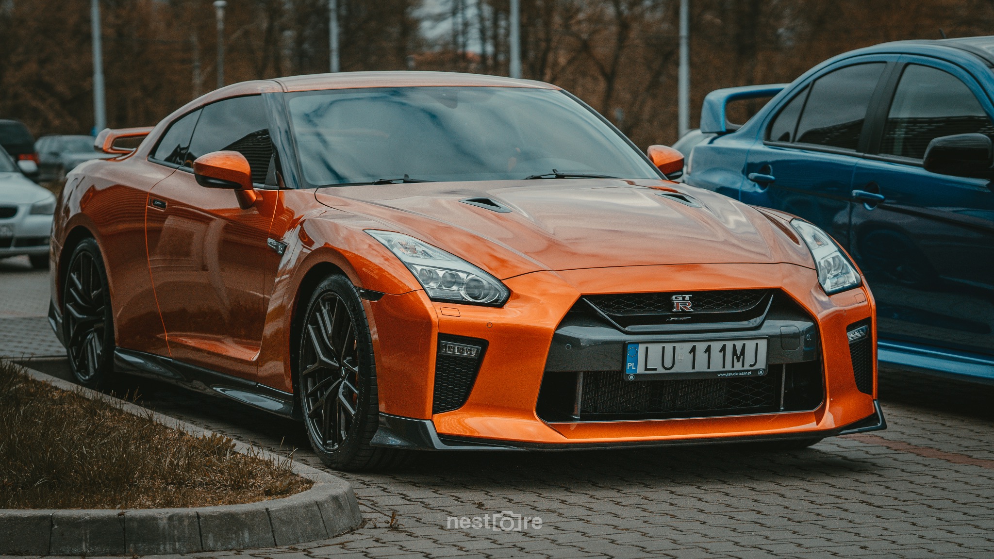Pomarańczowy Nissan GT-R na brukowanym parkingu, widok z przodu, tablica rejestracyjna LU 111MJ, obok zaparkowany niebieski samochód.