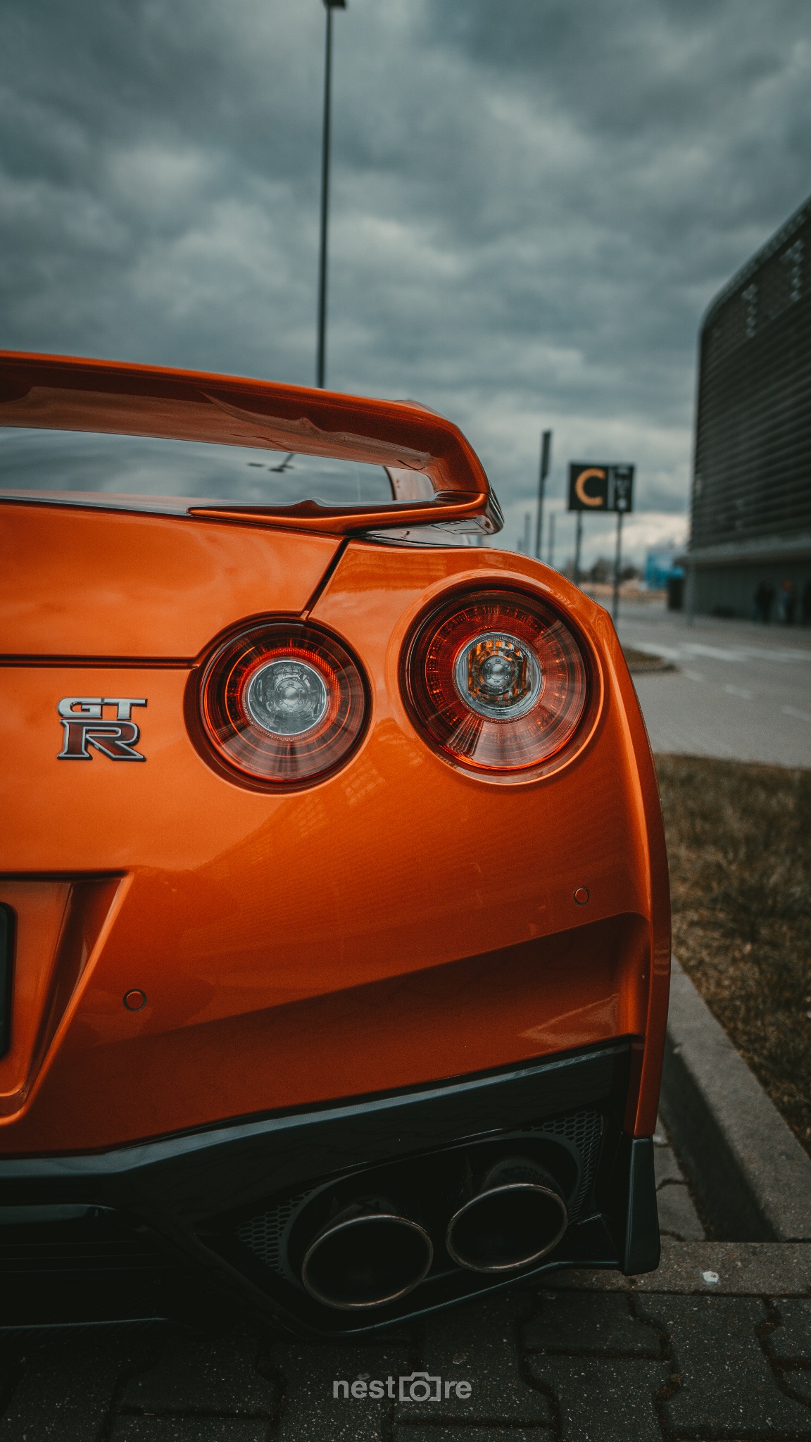 Pomarańczowy Nissan GT-R z widocznymi tylnymi światłami, logo GT-R i podwójnym wydechem, na tle pochmurnego nieba i budynku.