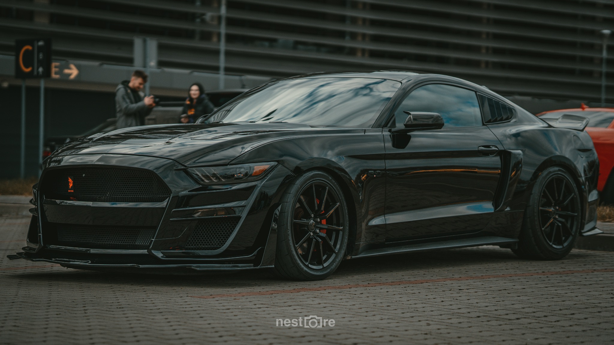 Czarny Ford Mustang Shelby GT500 na tle parkingu z dwoma osobami w tle, widoczne czerwone zaciski hamulcowe i logo na grillu.