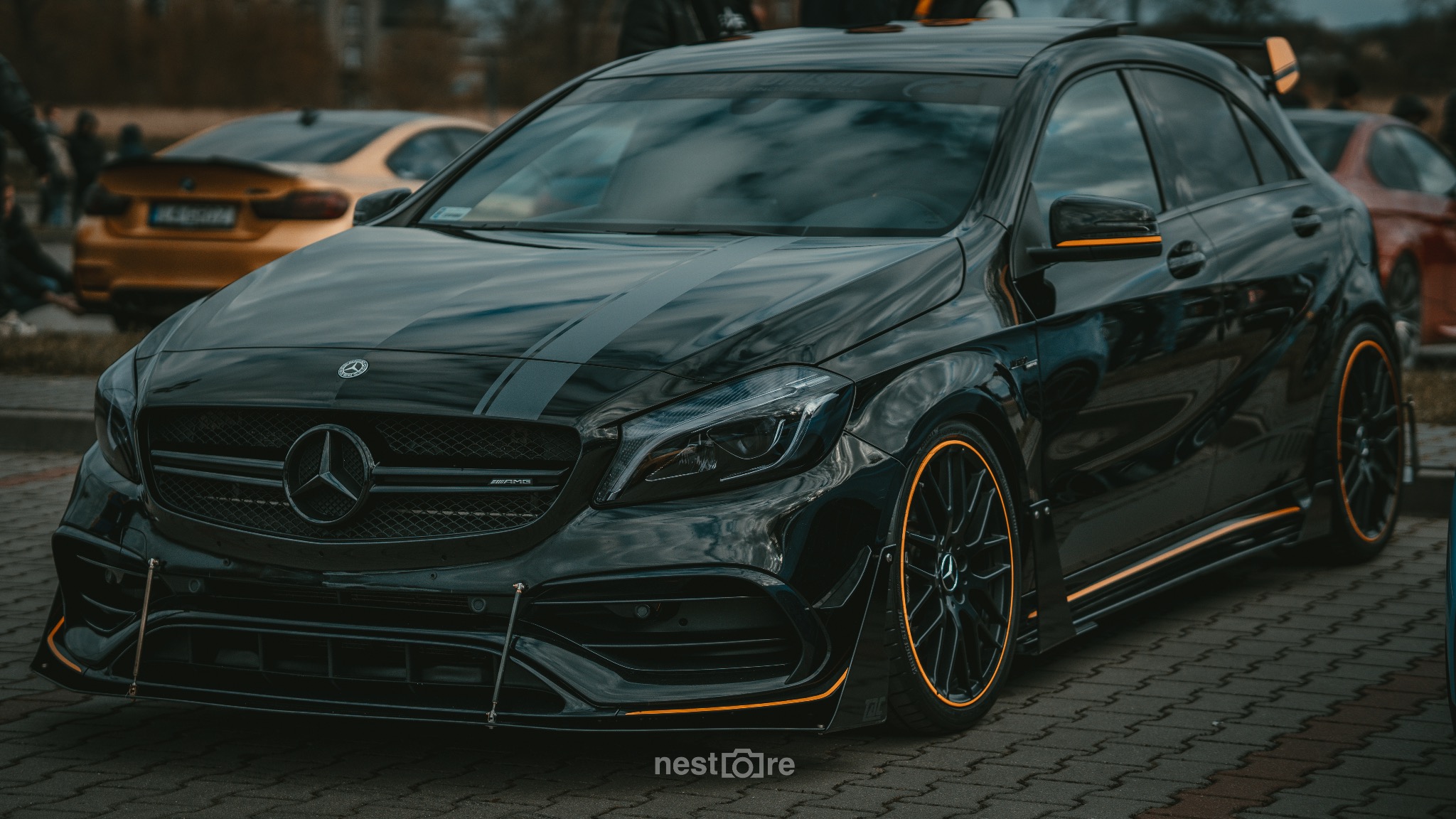 Czarny Mercedes-Benz A45 AMG z pomarańczowymi akcentami na felgach i listwach, zaparkowany na brukowanym placu, z innymi samochodami w tle.
