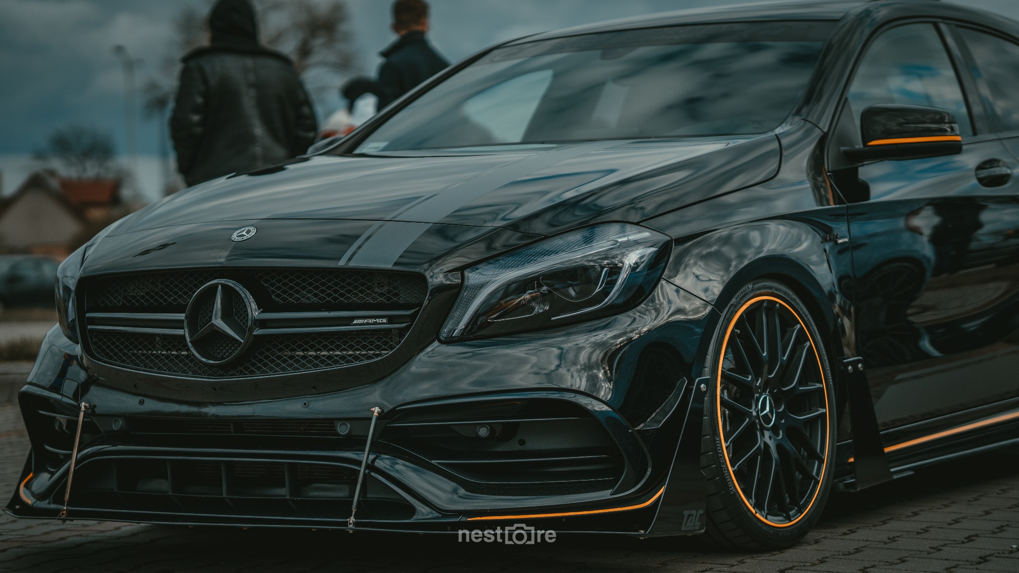 Czarny Mercedes A45 AMG z pomarańczowymi akcentami na felgach i lusterkach, widoczny grill z gwiazdą Mercedesa oraz logo AMG, na tle rozmyte sylwetki ludzi.
