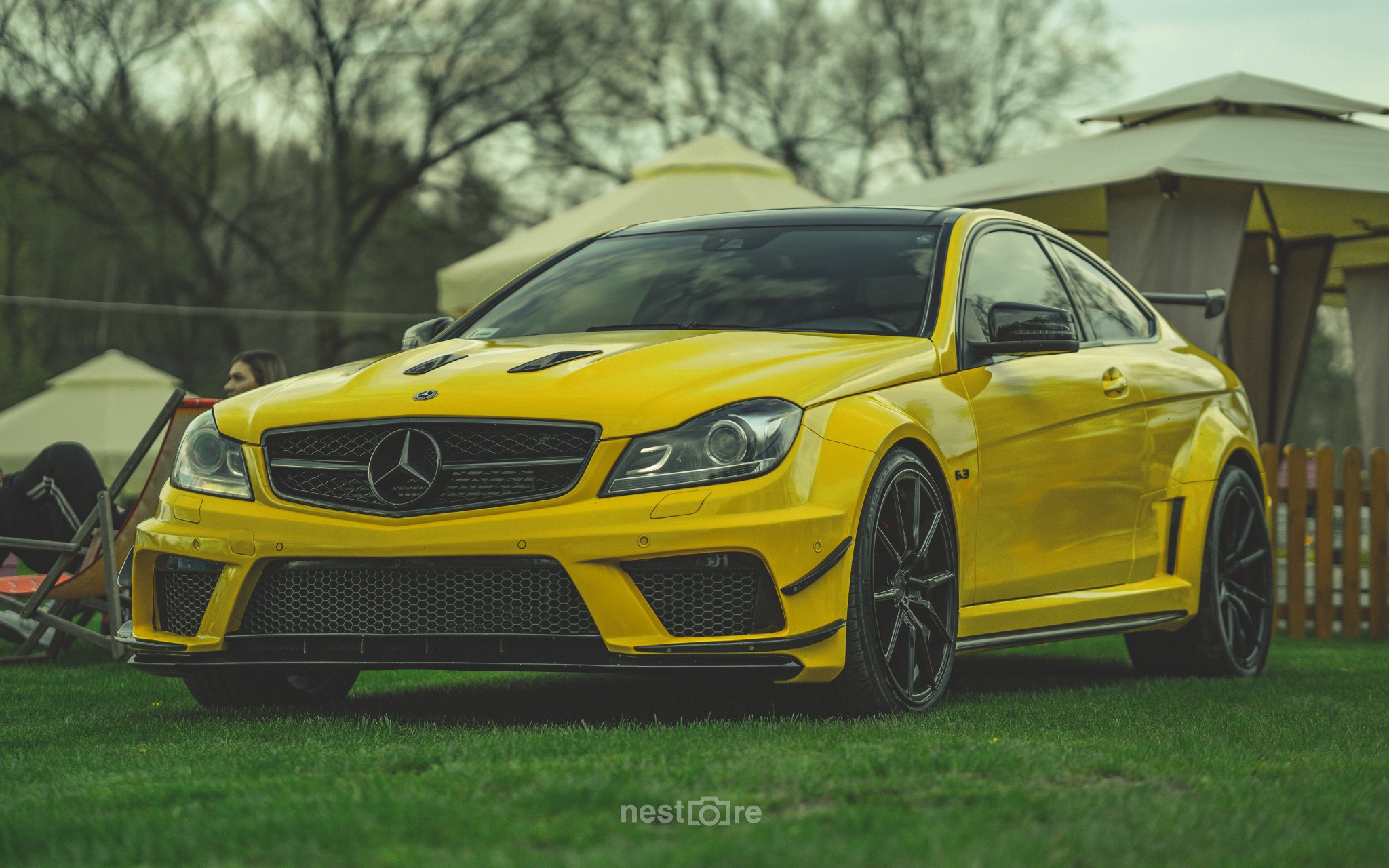 Żółty Mercedes C63 AMG Coupe na trawie, czarne felgi, tuningowany przód, w tle altana ogrodowa.