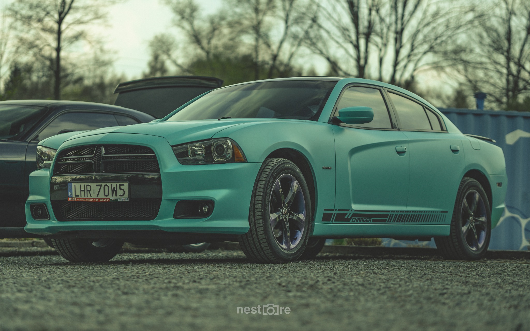 Turkusowy Dodge Charger zaparkowany na żwirowym parkingu, z widocznymi detalami jak czarne felgi i naklejki z napisem 'Charger' na boku.