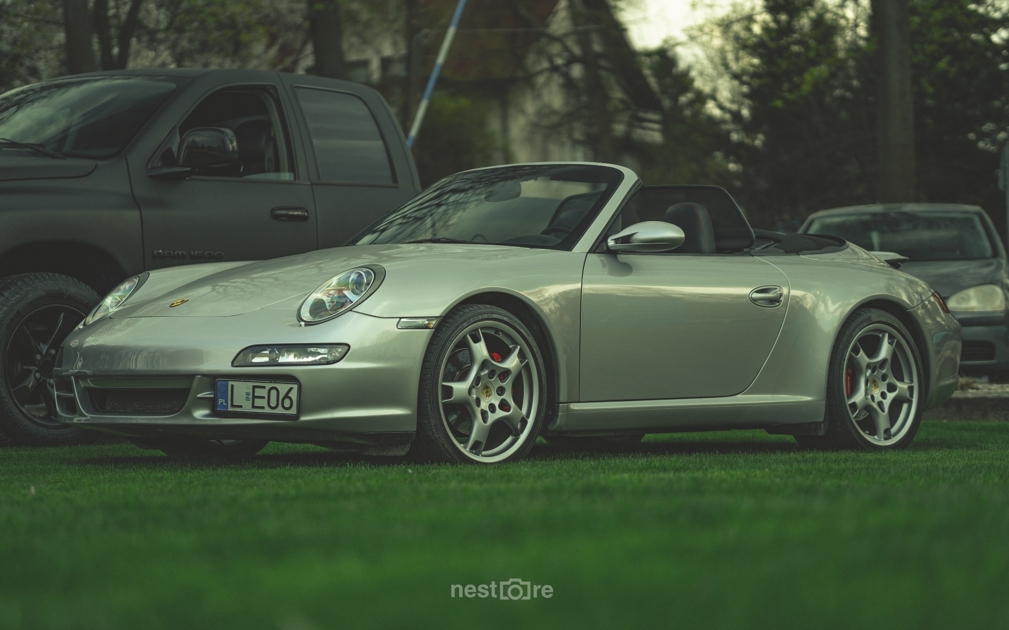 Srebrny Porsche 911 Cabriolet zaparkowany na trawie, z widoczną tablicą rejestracyjną i czerwonymi zaciskami hamulcowymi, w tle inne pojazdy.