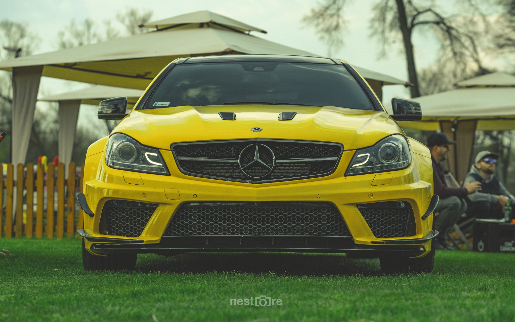 Żółty Mercedes C63 AMG stoi na trawie, widok z przodu, na tle altany i siedzących osób.