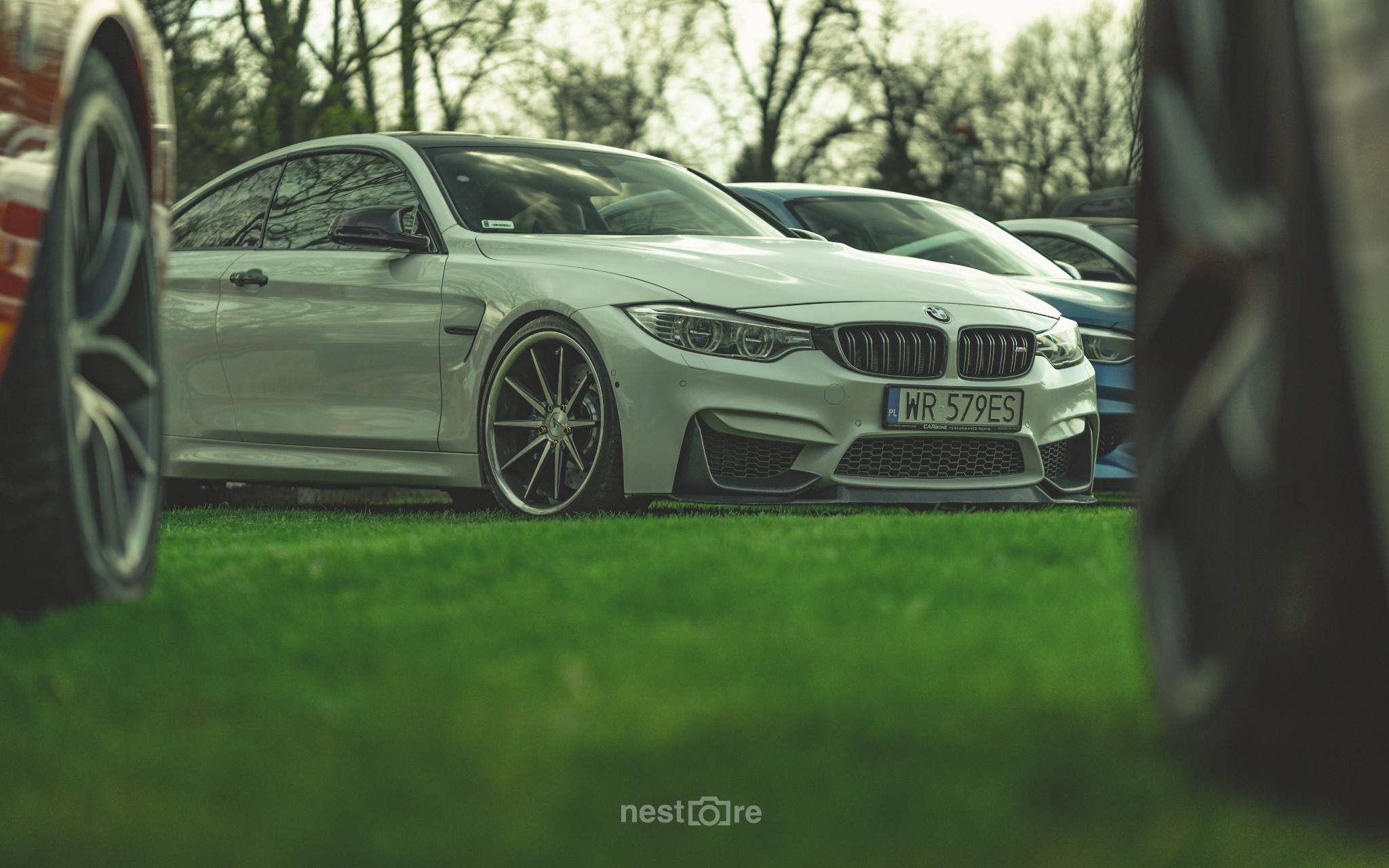 Jasnoszary samochód BMW M4 z polską tablicą rejestracyjną WR 579ES, zaparkowany na trawie, z widocznymi innymi samochodami w tle i fragmentami kół innych aut na pierwszym planie.