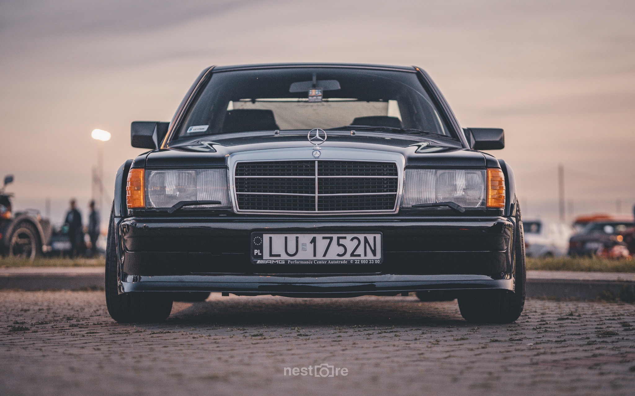 Czarny Mercedes-Benz W201 z Lublina, sfotografowany z przodu, z widoczną tablicą rejestracyjną LU-1752N i logo AMG na listwie pod tablicą, na tle parkingu o zmierzchu.