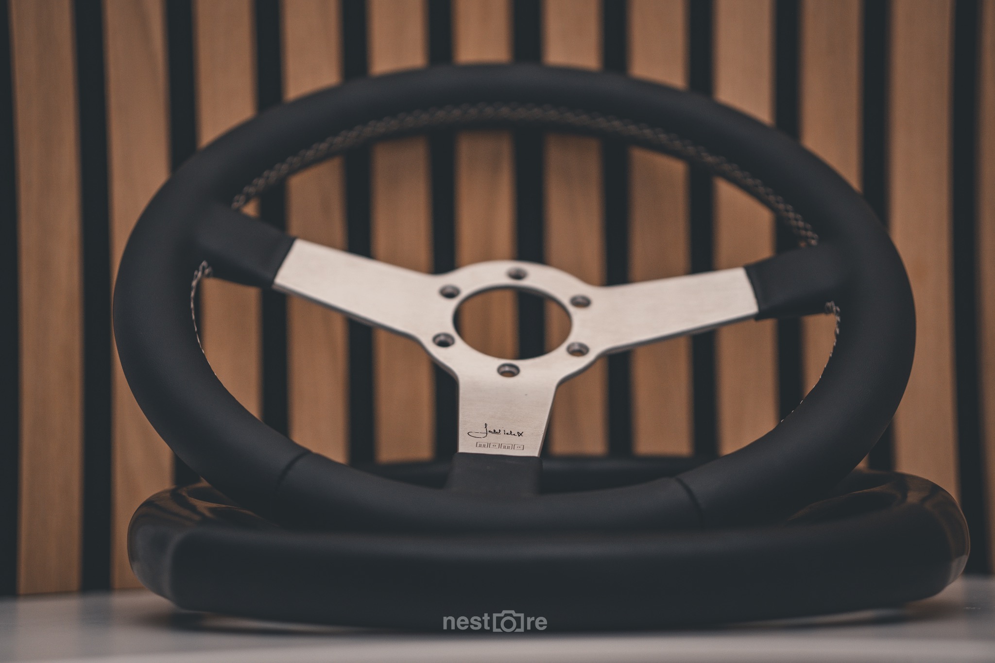 Dwa czarne skórzane koła kierownicy z metalowymi ramionami, jedno na drugim, z widocznym logo 'MOMO' na ramieniu, na tle brązowych pionowych desek.