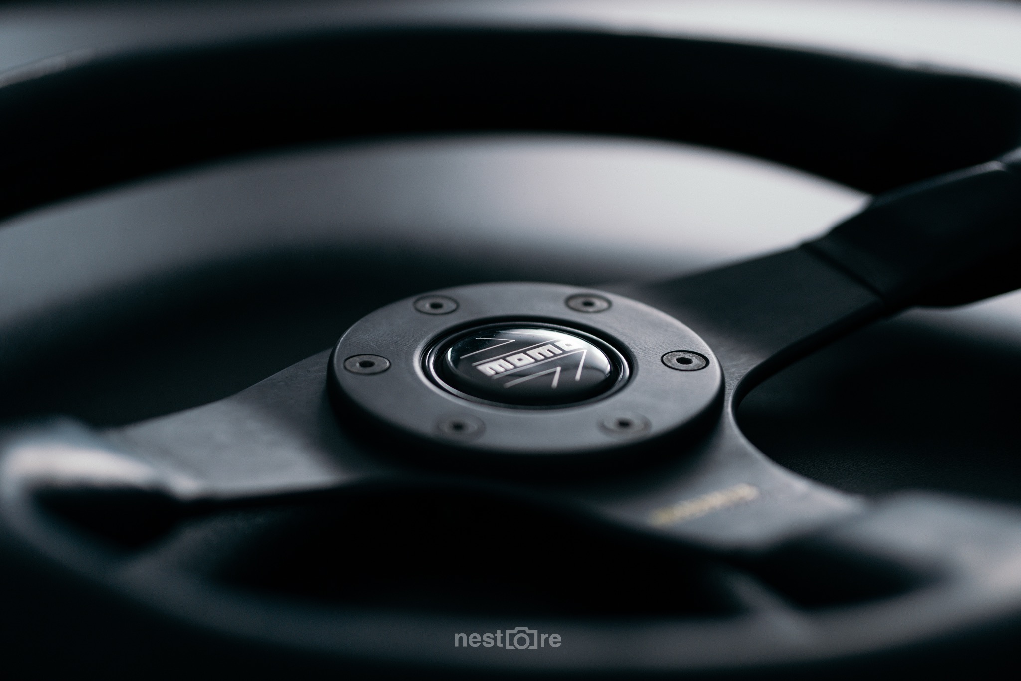 Czarna kierownica samochodowa MOMO z widocznym logo i śrubami mocującymi, sfotografowana z bliska w stonowanym oświetleniu.