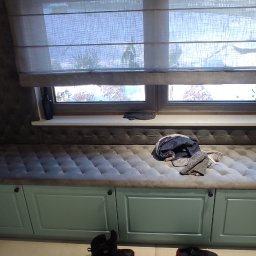 Dekor Max - Niski, turkusowy mebel z szufladami i tapicerowanym siedziskiem pod oknem z roletami rzymskimi, w przedpokoju z parasolami i kapciami.