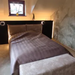 Sypialnia na poddaszu z ciemnym zagłówkiem z oświetleniem LED, wzorzystą tapetą z motywem tropikalnym i beżową tapicerowaną ławką.