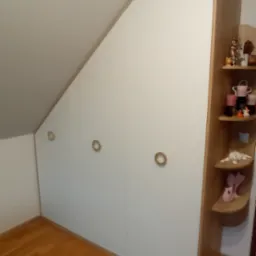 Biała szafa wnękowa z trzema drzwiami i okrągłymi uchwytami, dopasowana do skosu dachu, z drewnianymi półkami bocznymi, na których stoją ozdobne figurki, ustawiona na parkiecie.