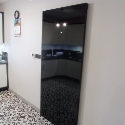 Dekor Max - Wysoka, czarna szafa z lustrzanym frontem w korytarzu, odbijająca wnętrze kuchni z białymi szafkami i czarnym blatem, wzorzysta podłoga w biało-czarne ornamenty.