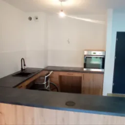 Nowoczesna kuchnia z blatem w odcieniach szarości i drewnianymi frontami, przystosowana dla osoby niepełnosprawnej poruszającej się na wózku inwalidzkim, widoczna czarna bateria i zlewozmywak.