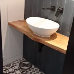 Biała, owalna umywalka nablatowa na blacie z jasnego drewna o nieregularnej krawędzi, z czarną armaturą i odpływem na tle ciemnoszarych płytek imitujących drewno i szafki z białymi frontami.