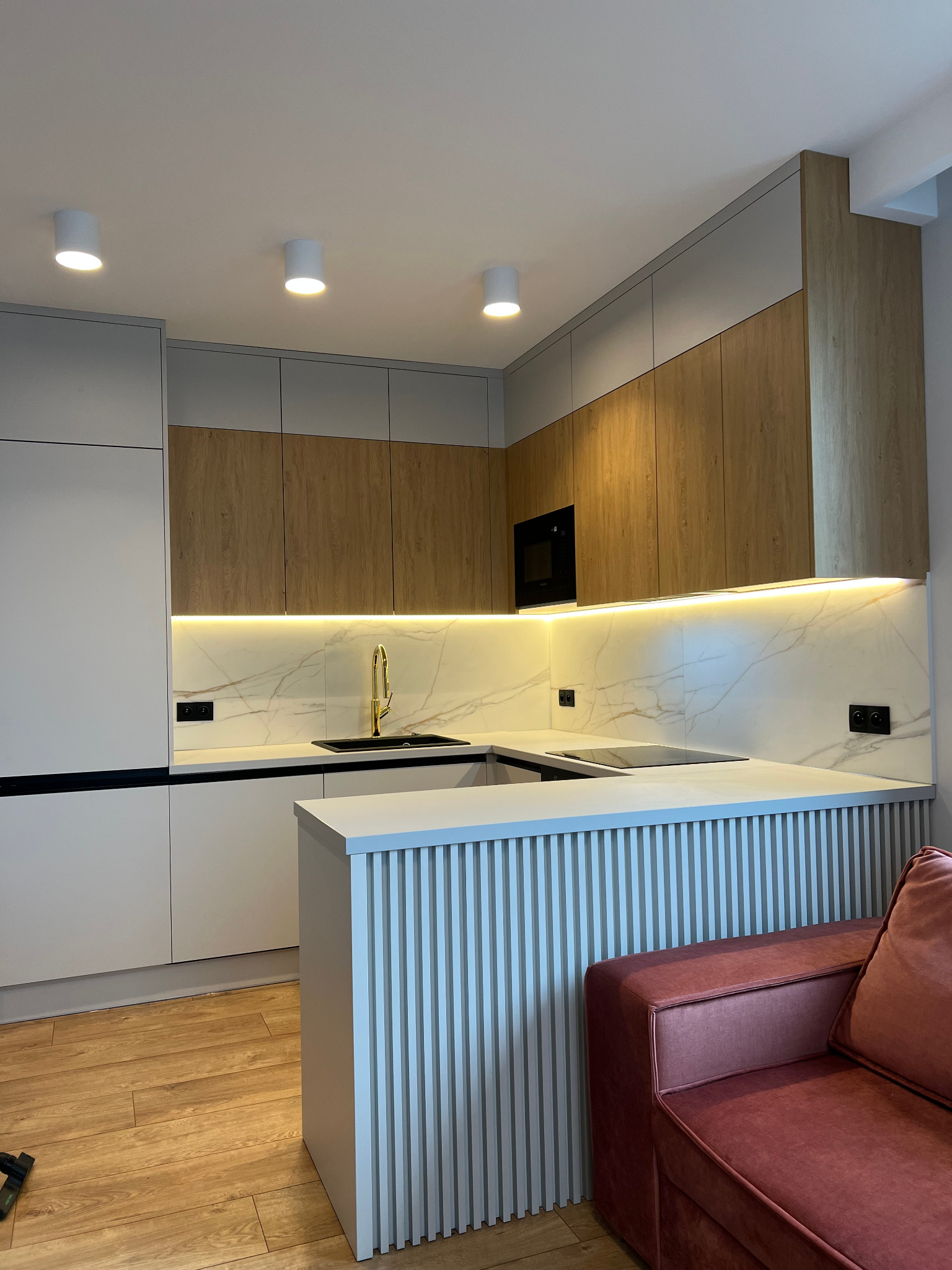 Nowoczesna kuchnia z frontami w kolorze drewna i szarości, oświetleniem LED podszafkowym, złotą baterią i wyspą z pionowymi listwami, widoczny fragment sofy w kolorze bordo.