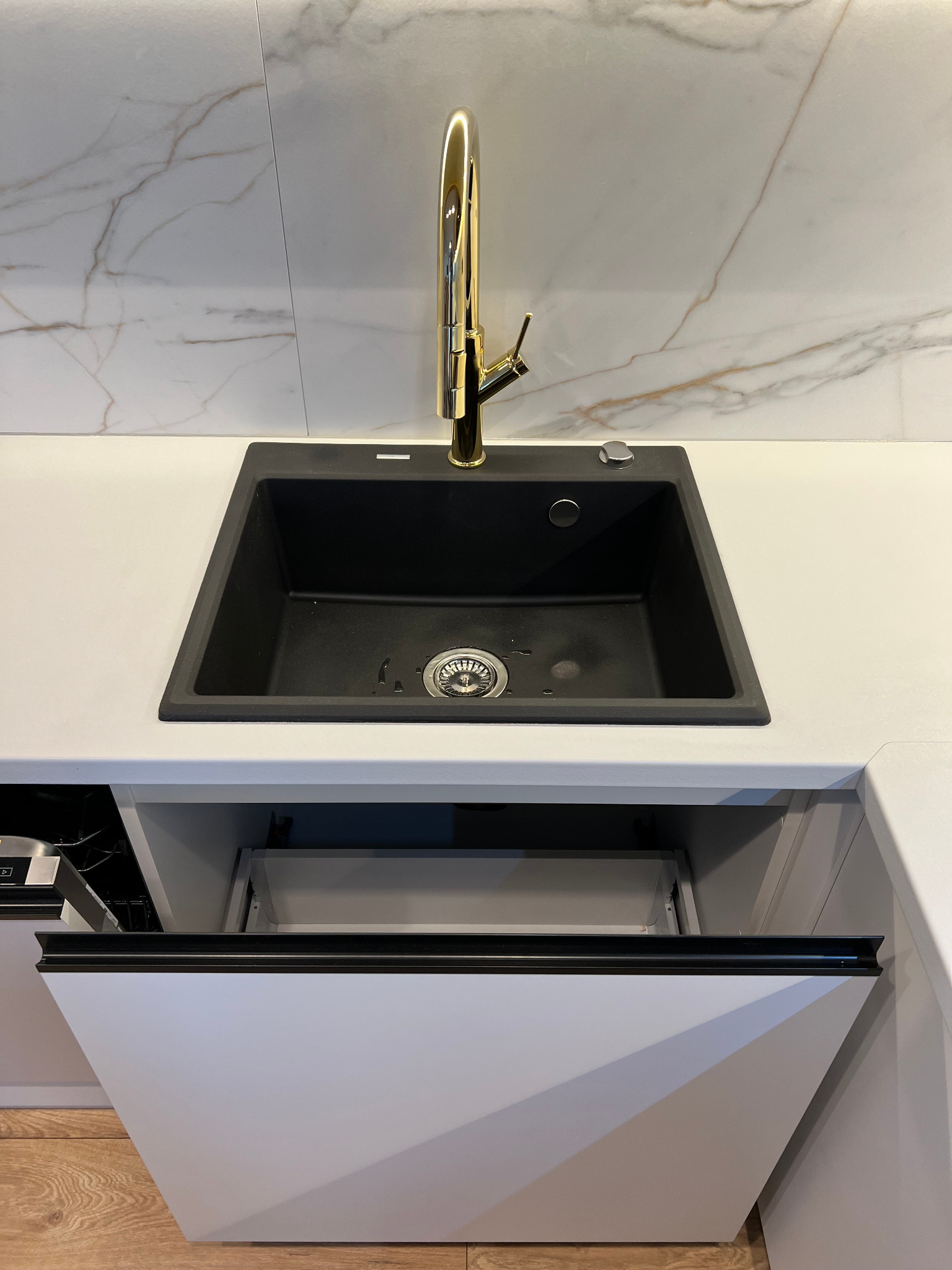 Czarny zlewozmywak granitowy z złotą baterią na blacie z otwartą szafką pod zlewem, marmurowe płytki na ścianie.