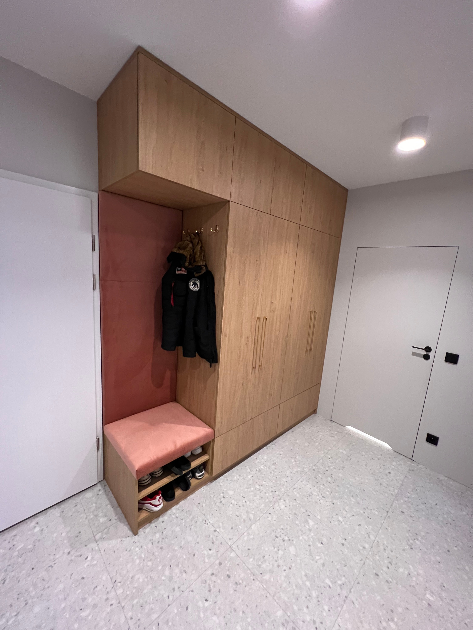Zabudowa przedpokoju z drewnopodobnymi frontami, siedziskiem obitym różowym welurem i otwartą półką na buty; na wieszakach wisi kurtka.