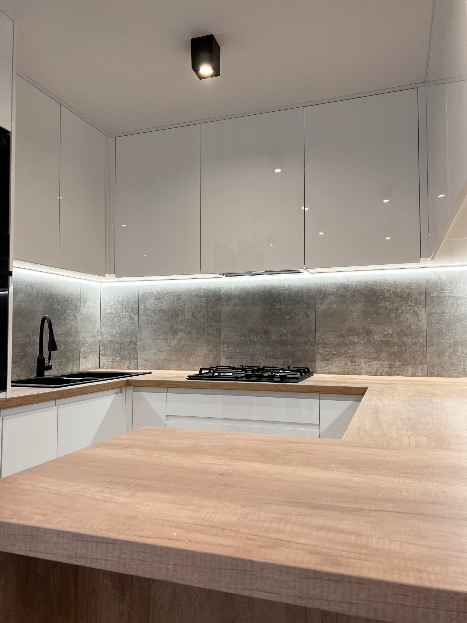 Nowoczesna kuchnia z białymi, lakierowanymi szafkami, drewnianym blatem, czarną armaturą i oświetleniem LED pod szafkami wiszącymi.