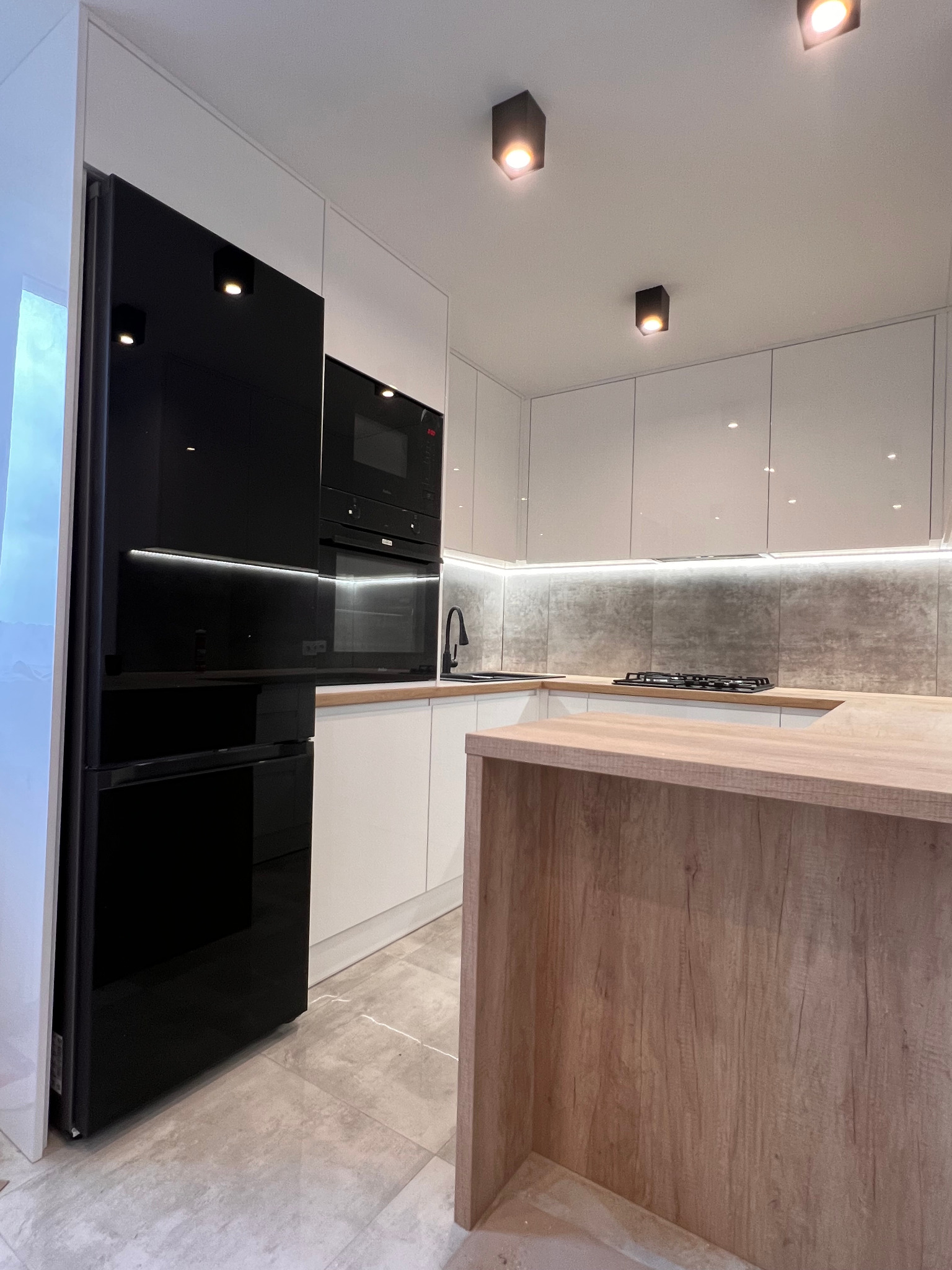 Nowoczesna kuchnia z białymi, błyszczącymi szafkami, czarnym sprzętem AGD (lodówka i piekarnik) oraz wyspą kuchenną z drewnianym blatem i wykończeniem imitującym drewno.