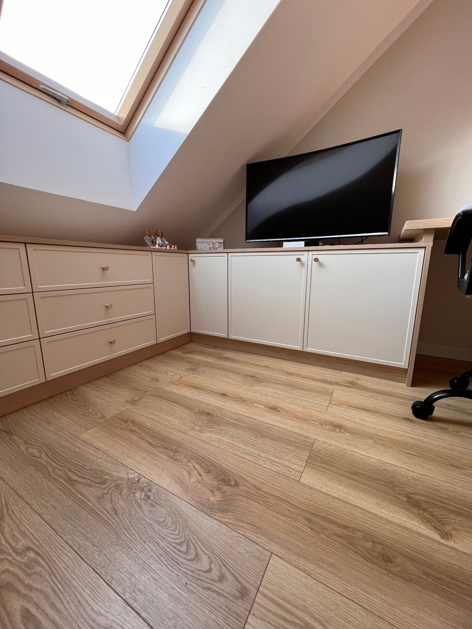 Zabudowa pod skosem na poddaszu z szafkami, szufladami z okrągłymi uchwytami i blatem pod telewizor, uzupełniona o drewnianą podłogę i widoczne okno dachowe.