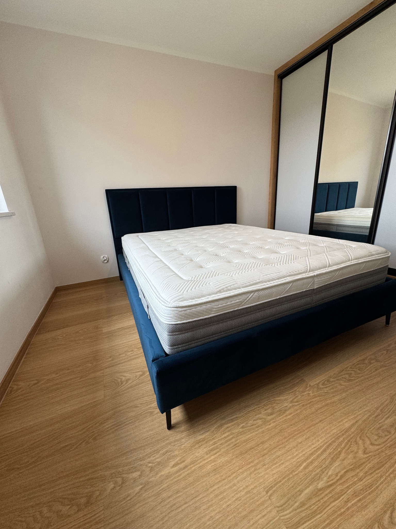 Nowoczesne łóżko z ciemnoniebieskim, tapicerowanym zagłówkiem i białym materacem w minimalistycznej sypialni z drewnianą podłogą i szafą z lustrzanymi drzwiami przesuwnymi.