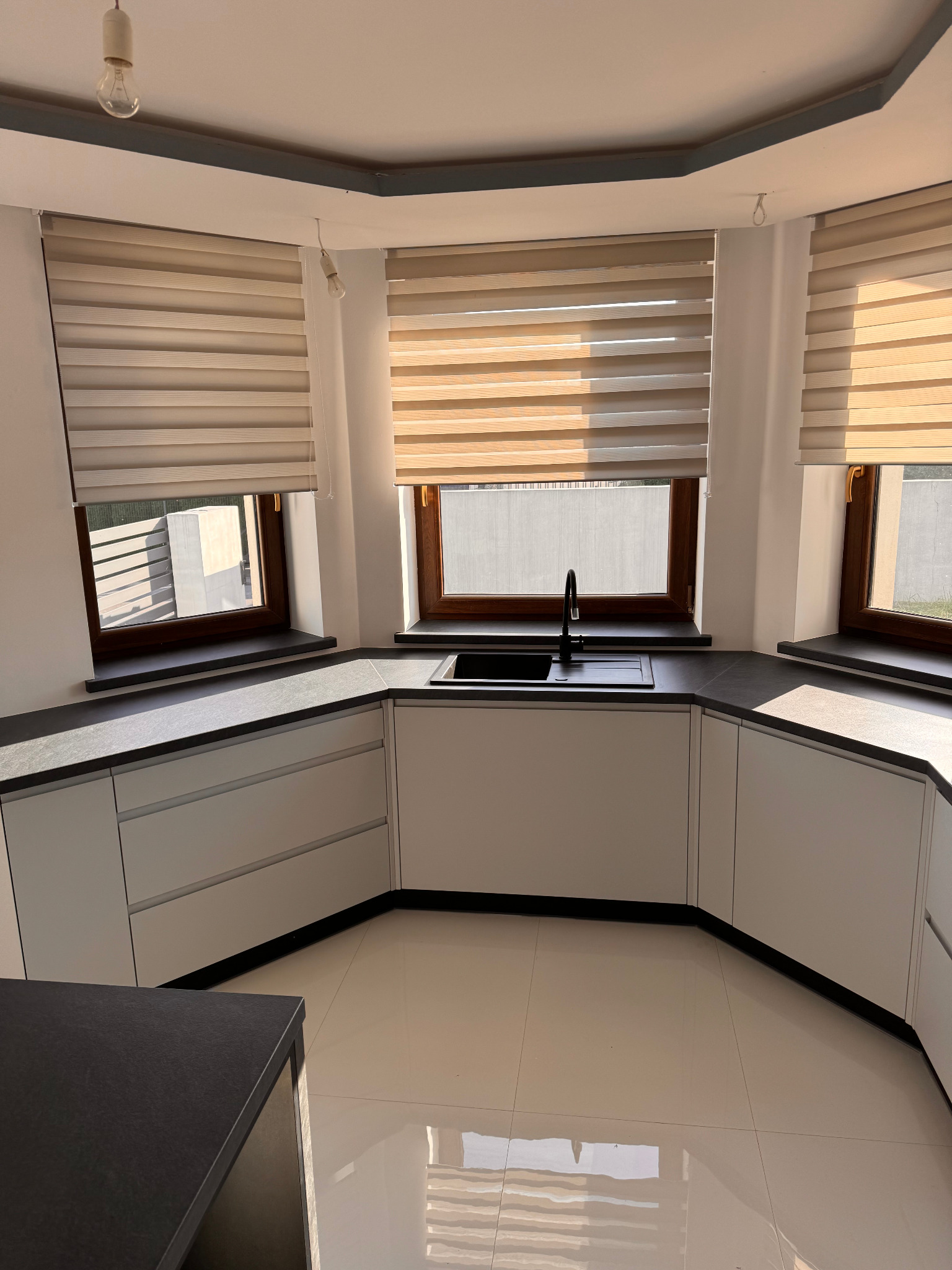 Jasna kuchnia z białymi szafkami i ciemnym blatem, okna z roletami dzień-noc, widok na zewnątrz, połyskująca podłoga.