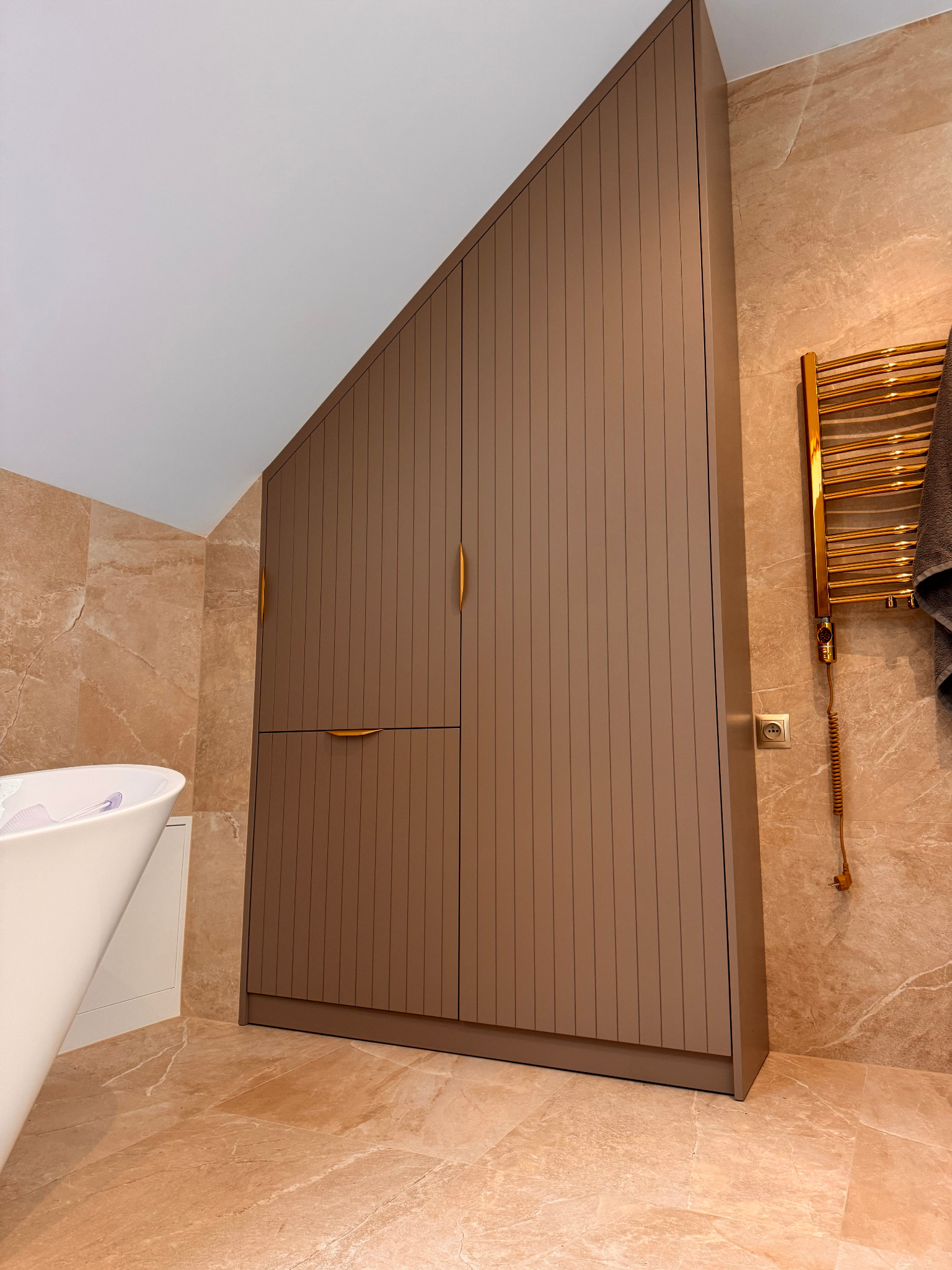Brązowa szafa wnękowa z pionowymi panelami i złotymi uchwytami w łazience wykończonej marmurem, obok wanny i grzejnika ręcznikowego.
