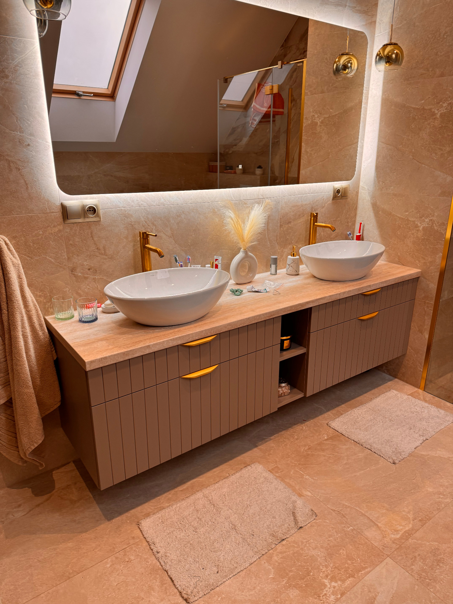 Łazienka w beżowych tonacjach z dwoma umywalkami nablatowymi, złotymi bateriami i szafką z ryflowanymi frontami oraz otwartą półką. Duże lustro z podświetleniem LED i widoczne okno dachowe.