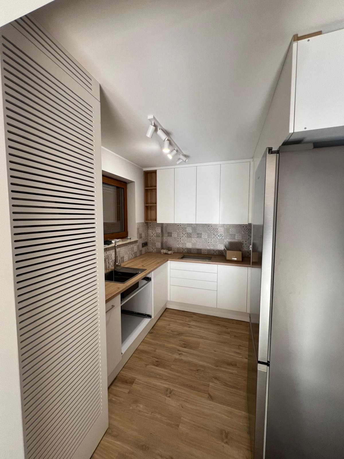 Nowoczesna kuchnia z białymi szafkami, drewnianym blatem, wzorzystą mozaiką na ścianie i szarymi lamelami na drzwiach po lewej stronie.