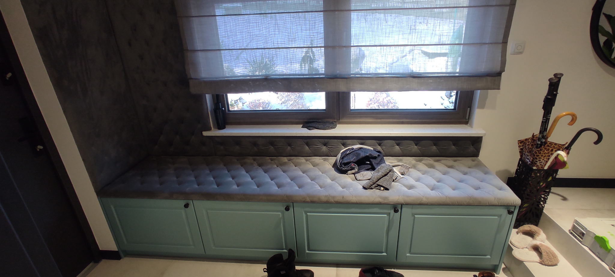 Niski, turkusowy mebel z szufladami i tapicerowanym siedziskiem pod oknem z roletami rzymskimi, w przedpokoju z parasolami i kapciami.